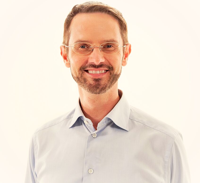 Dr. Markus Ebner, Portraitfoto. Er trägt ein eierschalen-farbenes Hemd, eine feine Brille sowie einen Dreitagebart. 