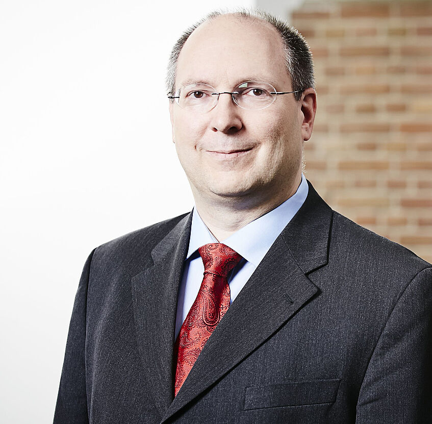 Portrait: Univ. Prof. Dr. Erik Hölzl. Er trägt einen Anzug mit roter Krawatte. 