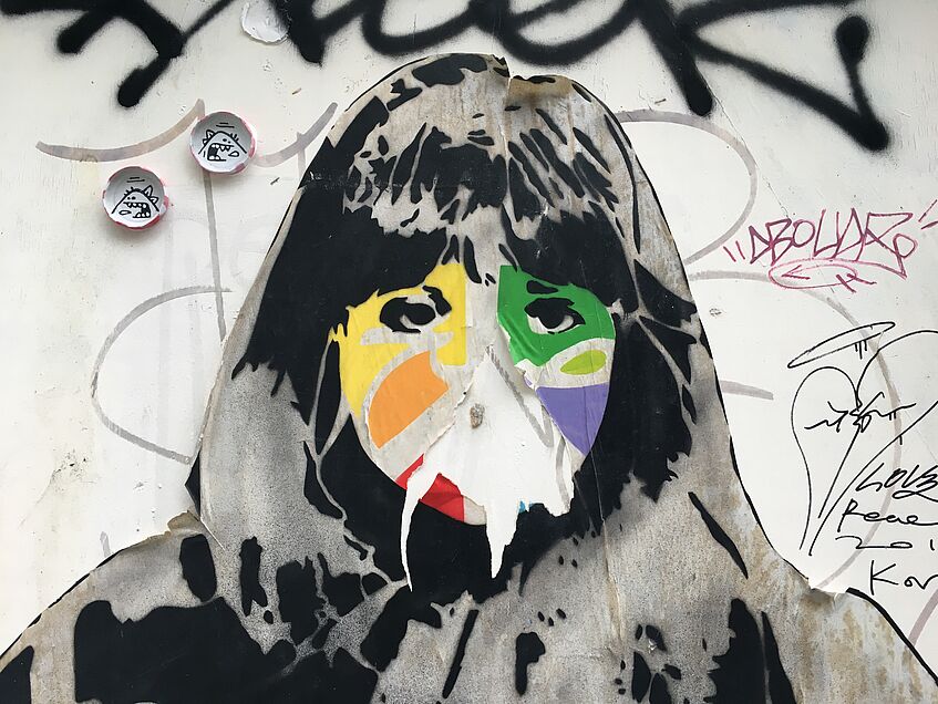 Straßenkunst. Der Kopf eines Mädchens, dargestellt mithilfe verschiedener Materialen wie Farbe sowie aufgeklebtem Papier. Umgeben von Graffiti. 
