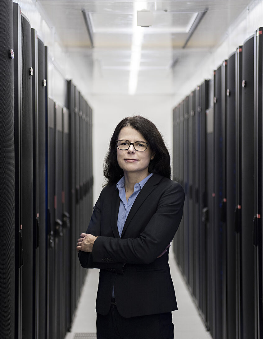 Dr. Petra Oswald-Ulreich steht mit verschränkten Armen in einem Flur, an dessen Seiten schwarze Spinde aufgereiht sind. Sie trägt eine blaue Bluse, schwarzen Anzug und eine schwarze Brille. Sie hat schwarze, lange Haare. 